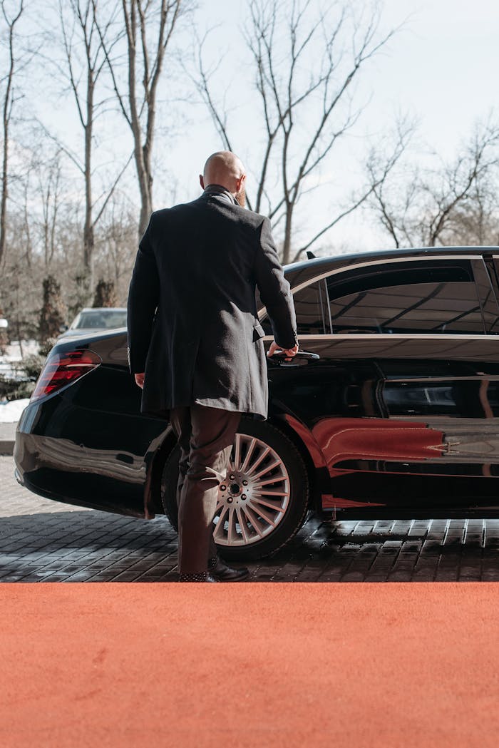 Bald man in a black suit approaches a élégant black car on a sunny day.
